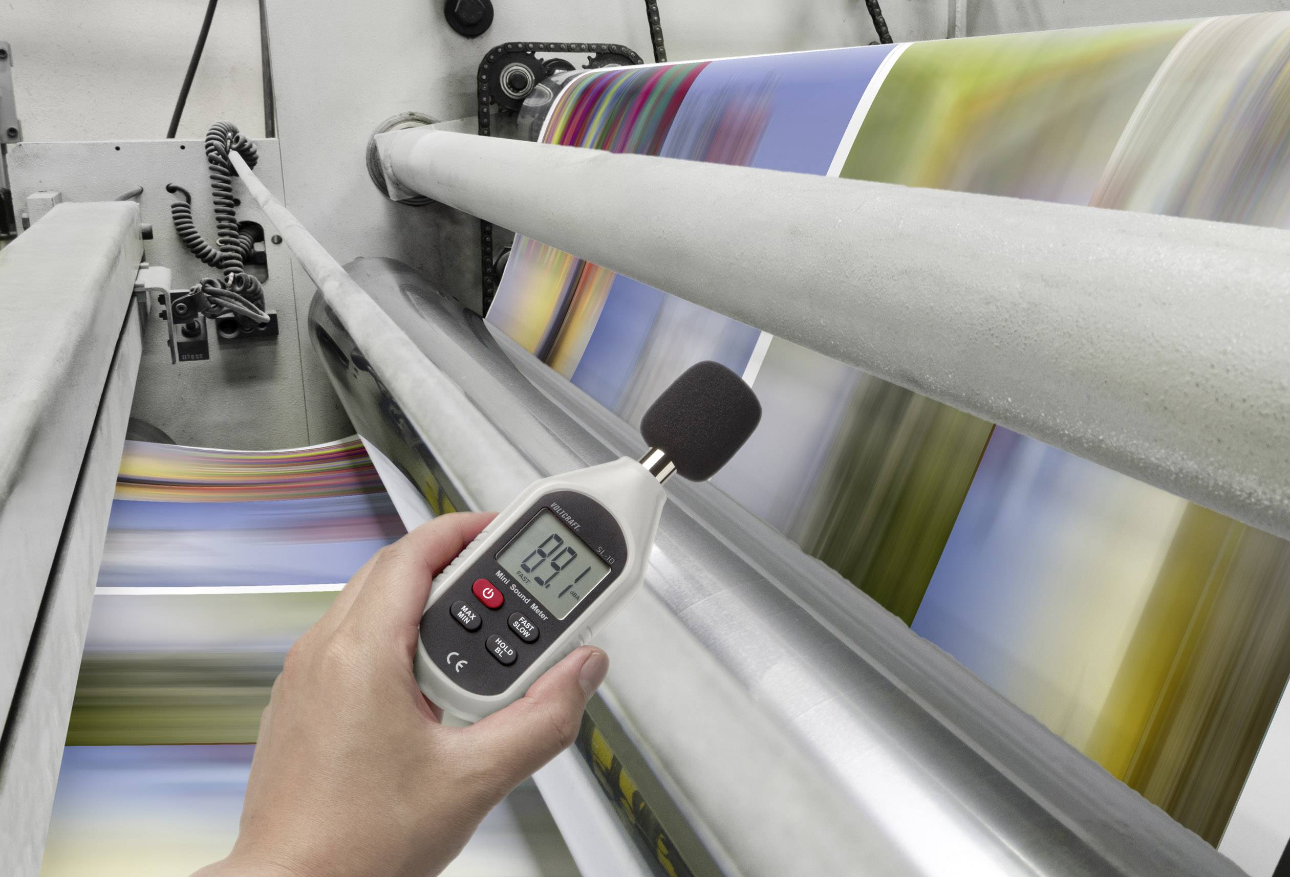 Eine Person misst mit einem Dezibelmeter den Lärmpegel einer Druckmaschine, die gerade bunte Seiten bedruckt.