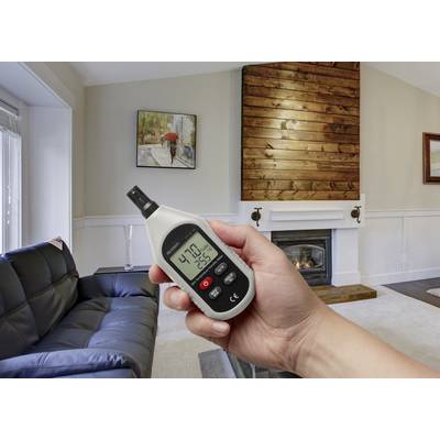 Eine Hand hält ein Hygrometer in einem Wohnzimmer, zeigt 47,0% Luftfeuchtigkeit und 25,6°C an, Hintergrund ein Kamin und Sofa.