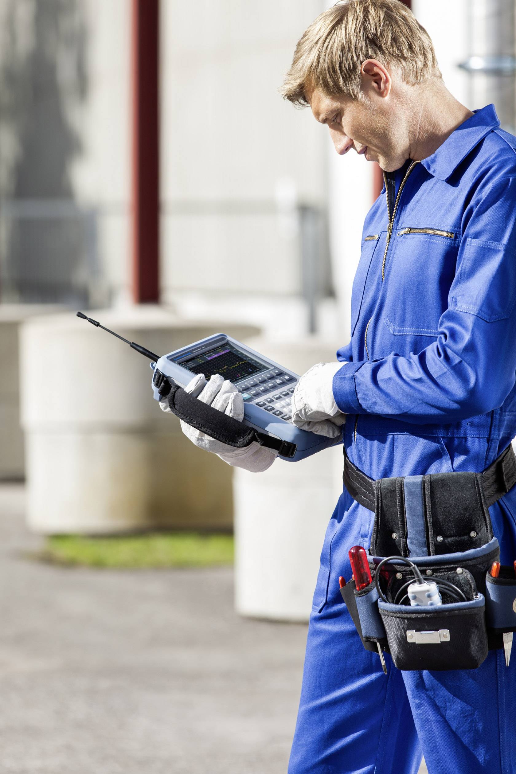 Ein Techniker in einem blauen Overall überprüft mit einem elektronischen Messgerät im Freien Daten oder Messungen.