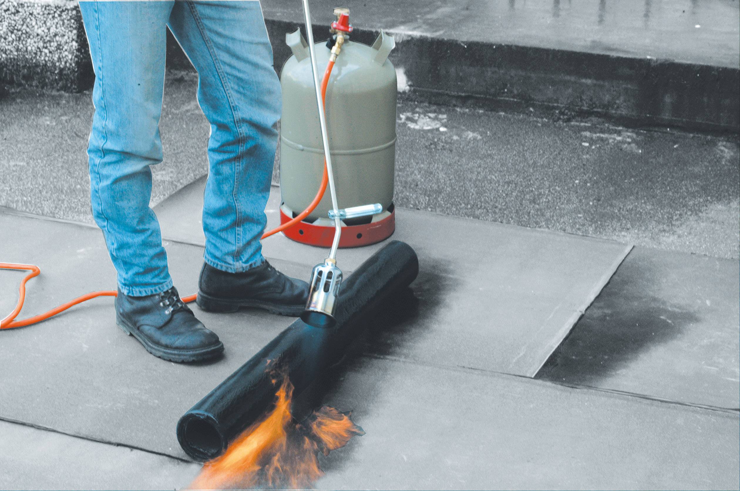 Person flext Bitumenbahn mit Gasbrenner auf Dachfläche. Gasflasche mit Schlauch verbunden. Wetterabdichtung bei Bauarbeiten.