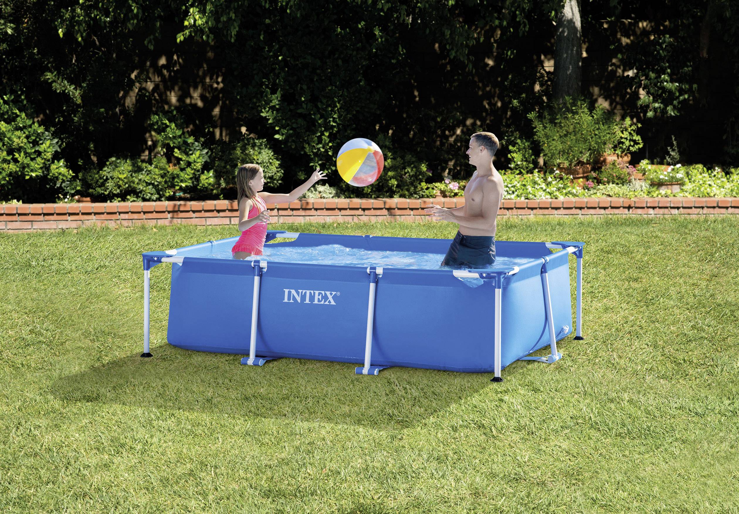 Zwei Personen spielen mit einem Ball in einem blauen rechteckigen Aufstellpool im Garten. Die Szenerie wirkt sommerlich und entspannt.
