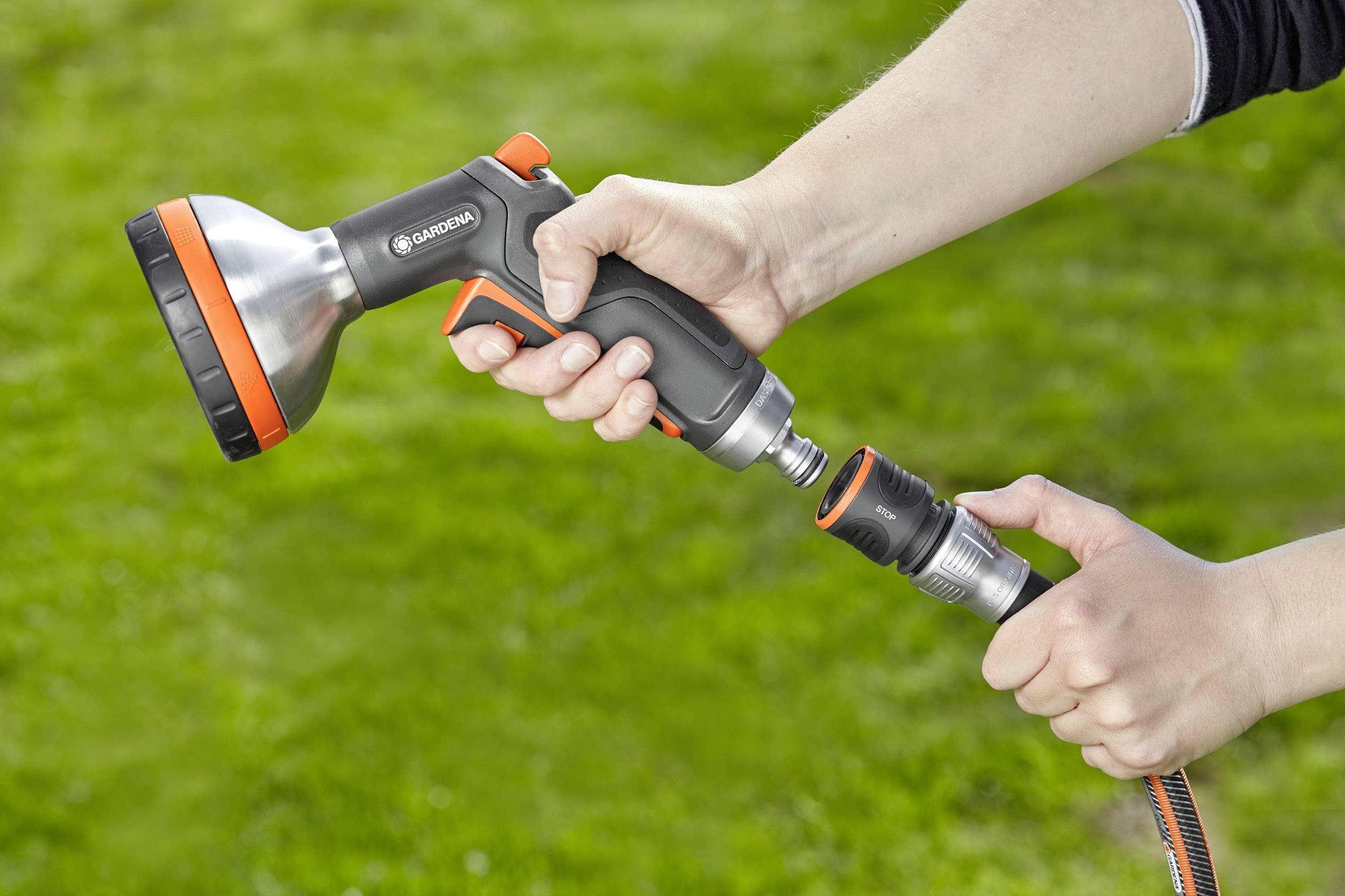 Eine Person hält eine Gardena-Gartenspritze und befestigt einen Schlauch daran. Im Hintergrund ist eine Rasenfläche zu sehen.