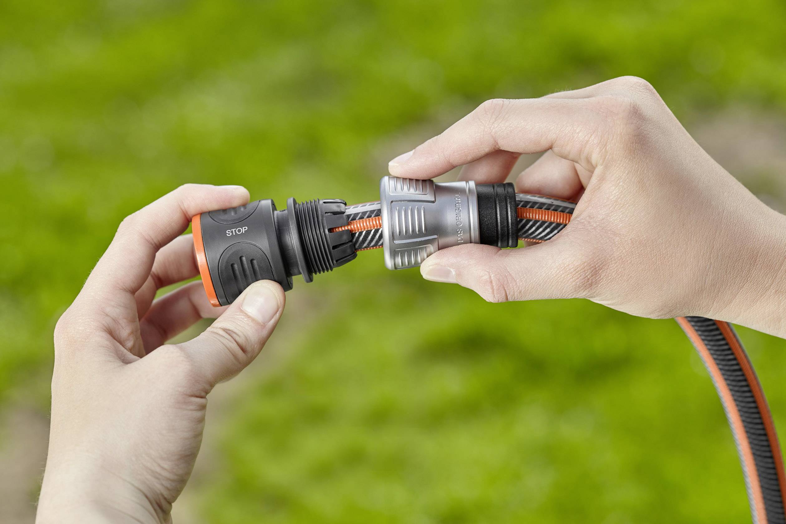 Zwei Hände verbinden zwei Gartenschläuche mit einem Klicksystem vor unscharfem grünem Hintergrund.