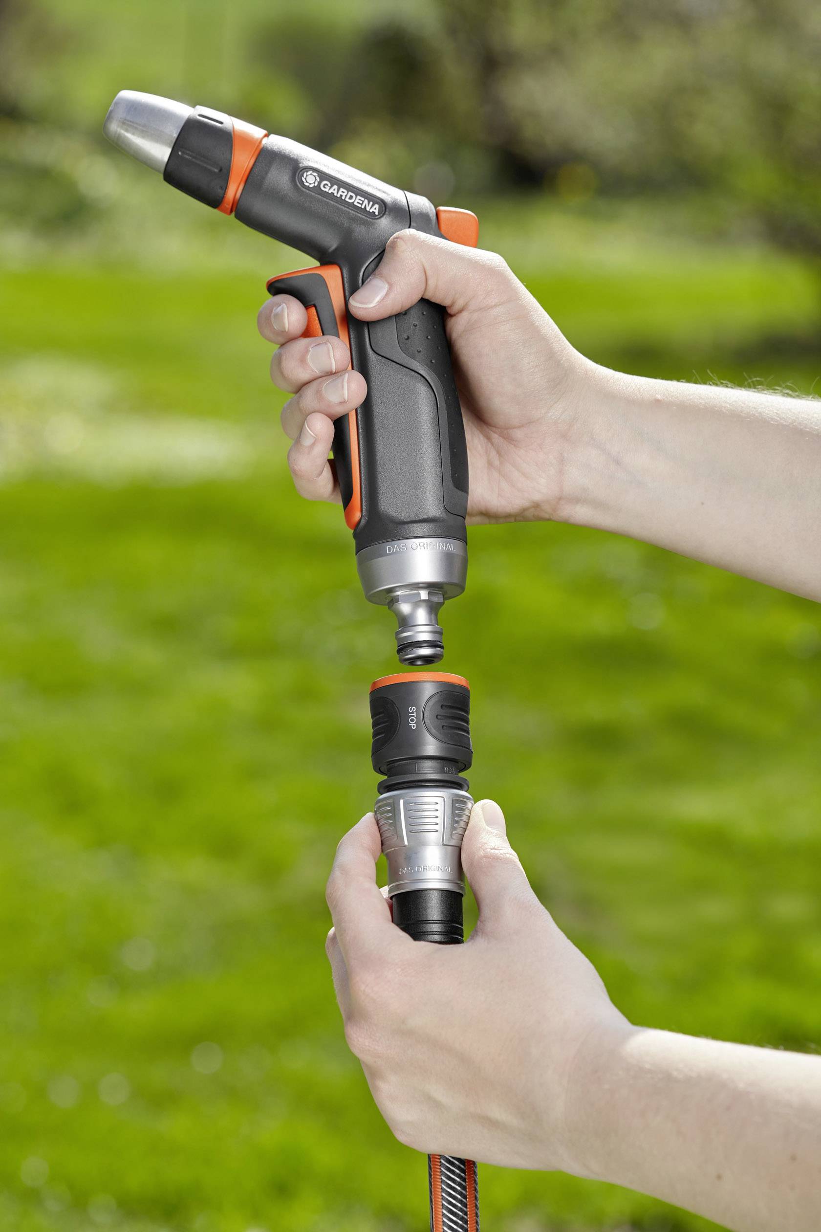 Person hält eine schwarze und orangefarbene Gartendüse, die an einen Wasserschlauch angeschlossen wird, vor einem grünen Rasen.