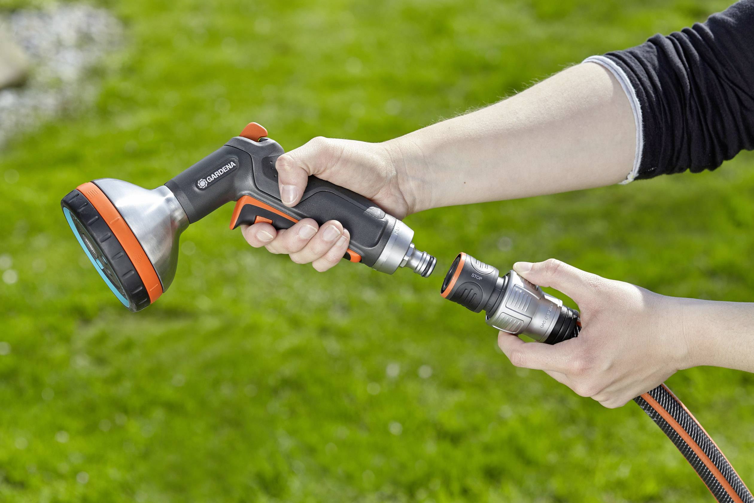 Person hält einen Gartenschlauchaufsatz und schließt ihn an einen Schlauch an, vor einem grünen Rasen im Freien.