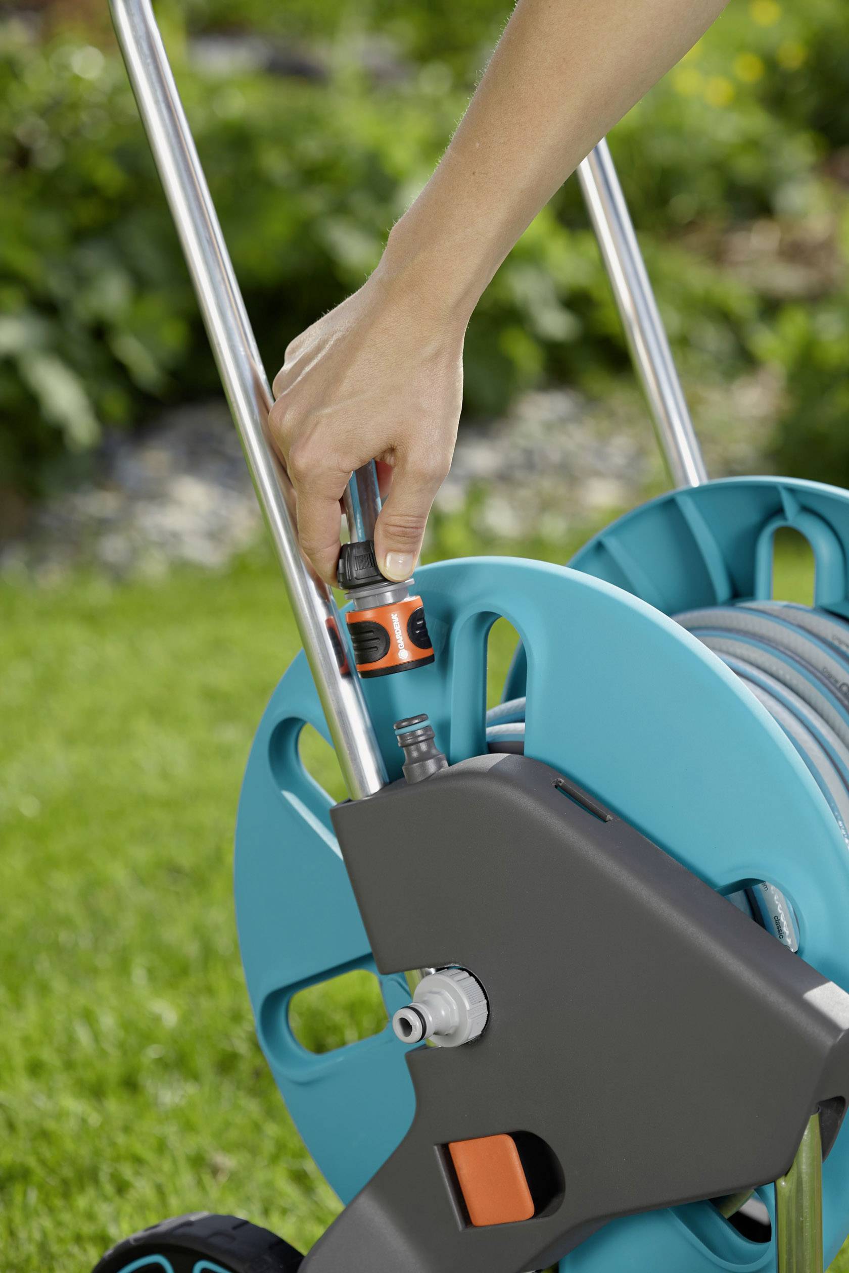 Eine Hand dreht den Wasseranschluss einer Gartenschlauchhaspel im Garten fest. Der Schlauchhalter ist auf Rasen abgestellt.