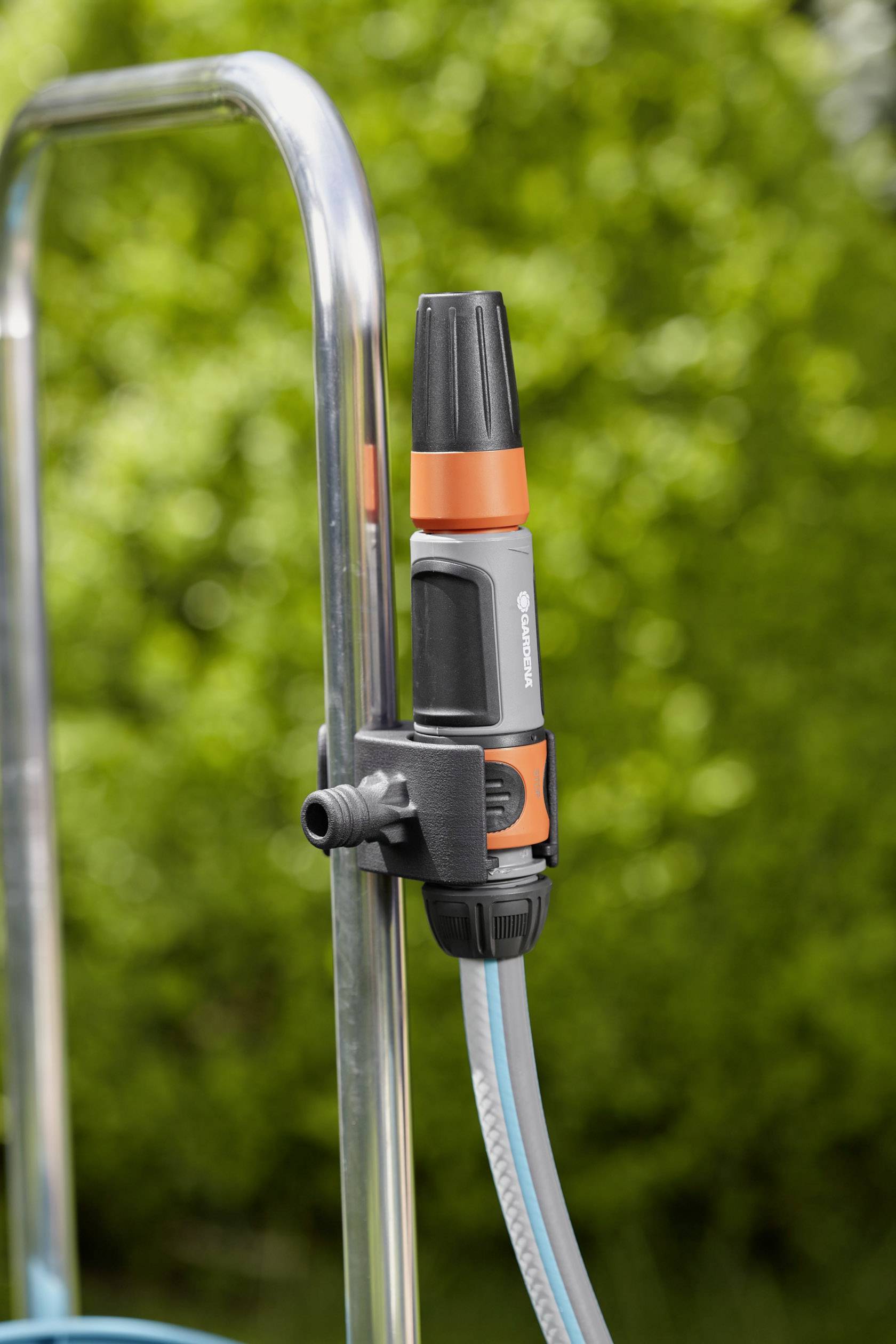 Ein Wasserschlauch mit orange-grauem Anschlussventil ist an einem Metallrahmen befestigt, vor unscharfem grünen Hintergrund.