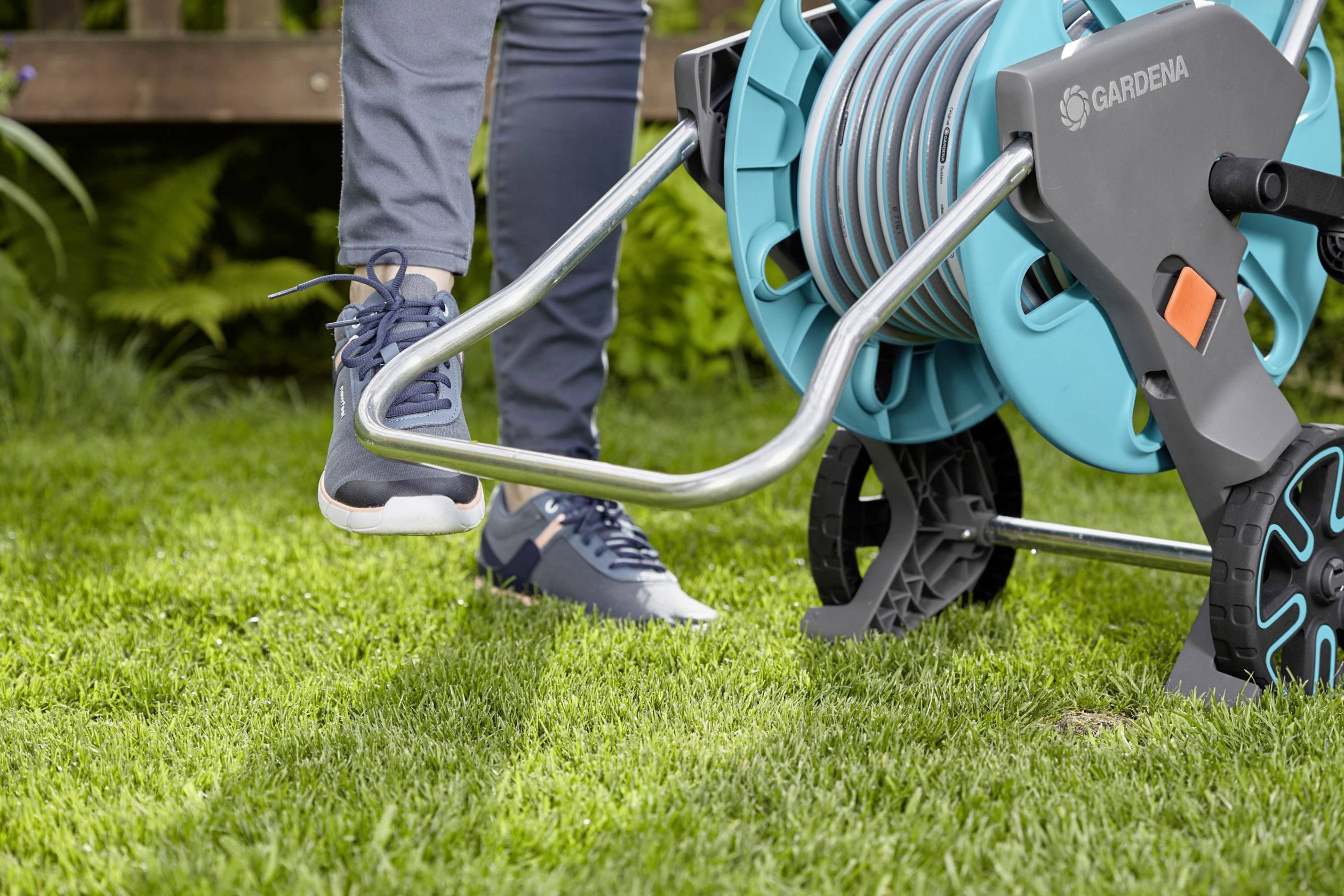 Person betätigt mit dem Fuß einen Gartenschlauchwagen auf einer Rasenfläche. Der Schlauch ist sauber aufgewickelt.