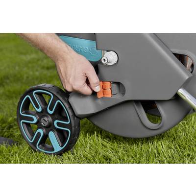 Eine Hand hält einen orangenen Hebel an einem Gartenschlauchwagen auf einer Rasenfläche. Der Schlauchwagen hat schwarze Räder mit blauen Akzenten.