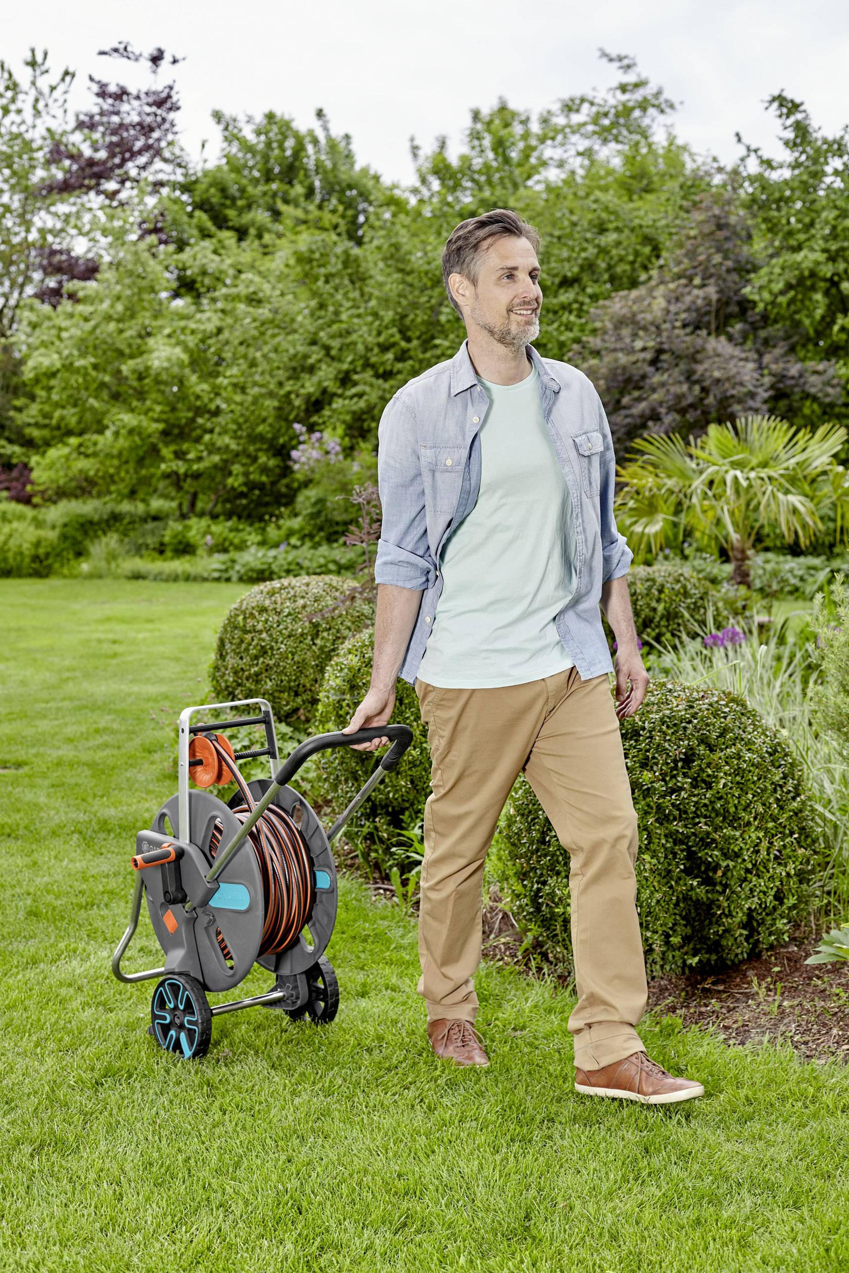 Ein Mann zieht einen Gartenschlauchwagen durch einen gepflegten Garten. Im Hintergrund sind Büsche und Bäume sichtbar.