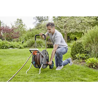 Ein Mann kniet auf einer Rasenfläche und rollt einen Gartenschlauch auf einen Schlauchwagen auf. Im Hintergrund sind Bäume und Sträucher.