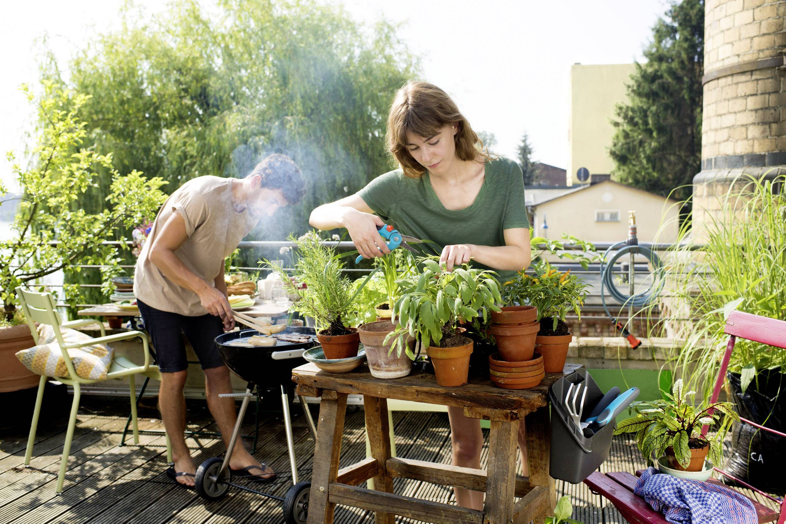Eine Frau gießt Pflanzen auf einem Tisch, während ein Mann im Hintergrund am Grill Essen zubereitet. Es ist eine sonnige Terrasse.