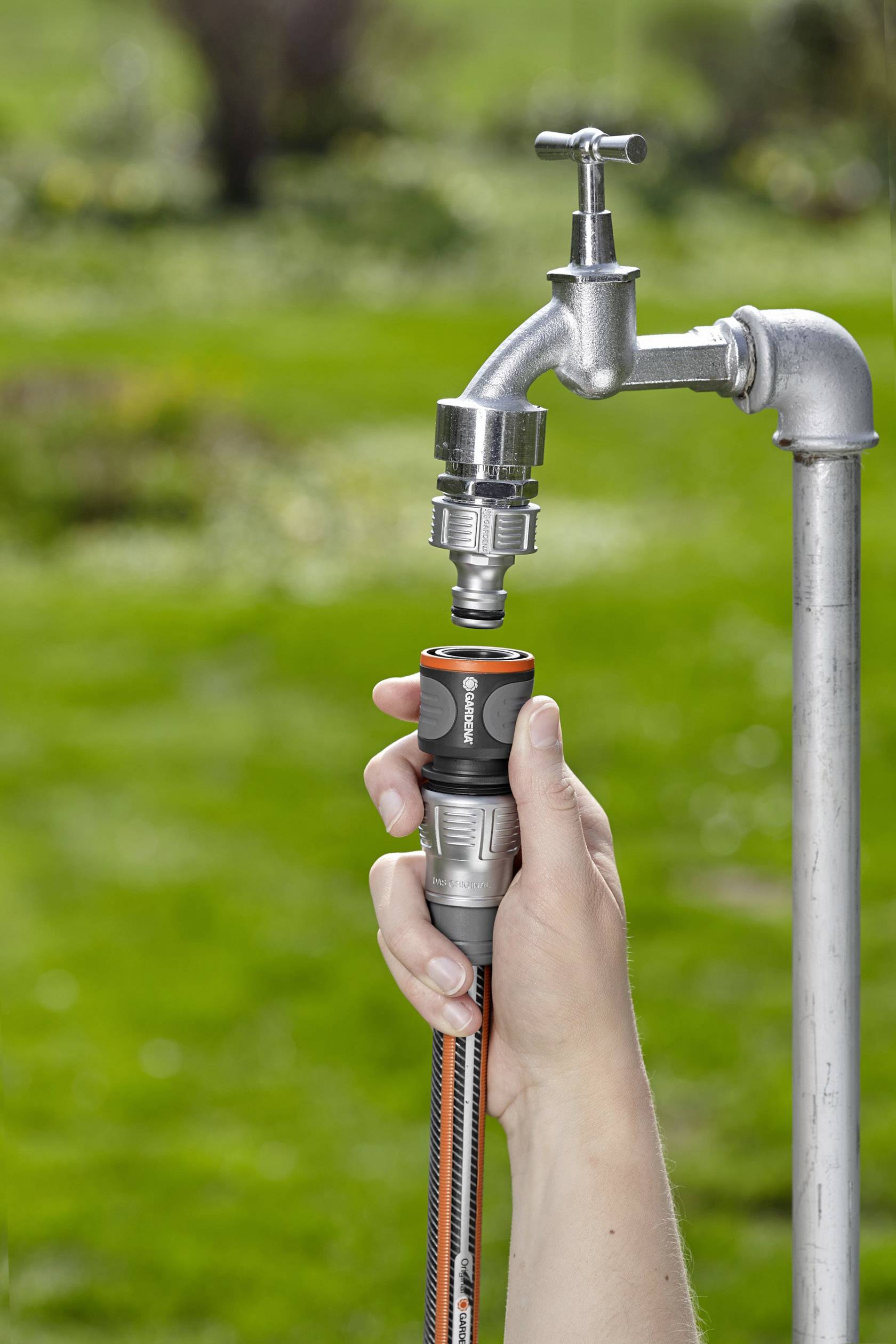 Eine Hand steckt einen Gartenschlauchanschluss an einen Wasserhahn im Freien. Im Hintergrund ist eine grüne Rasenfläche zu sehen.