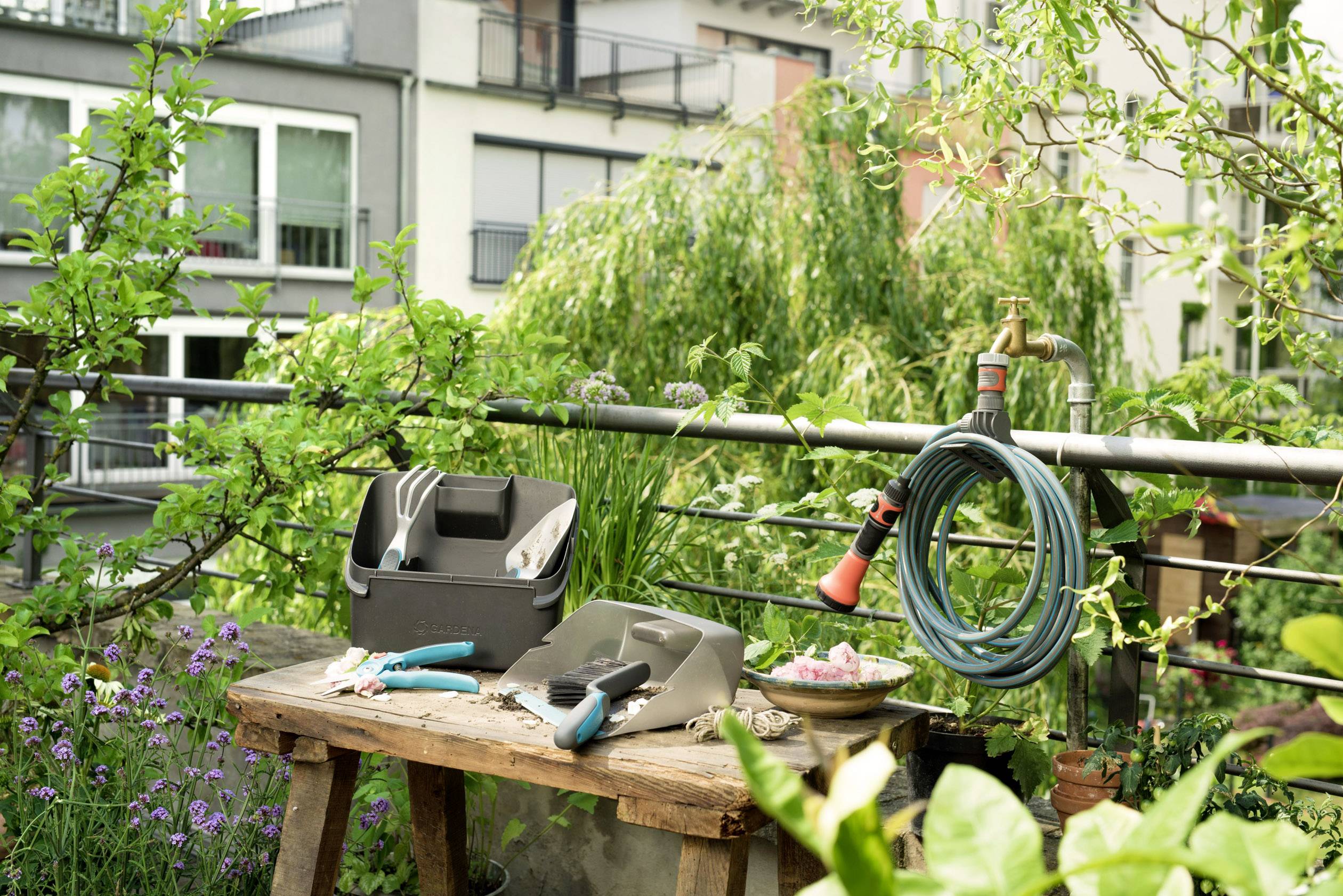 Ein Holztisch im Garten mit Gartenwerkzeugen, einer Gießkanne und einem Wasserschlauch vor einem Wohngebäude im Hintergrund.