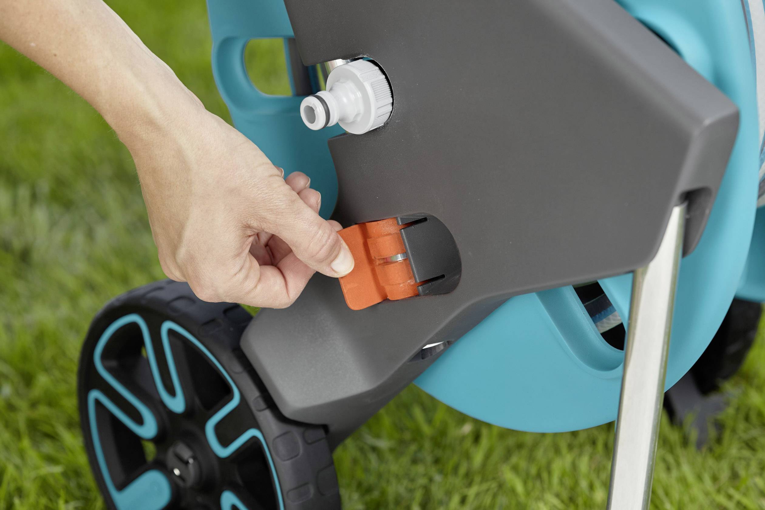 Eine Hand schaltet einen orangefarbenen Hebel an einem blauen Gartenschlauchwagen auf grüner Wiese.