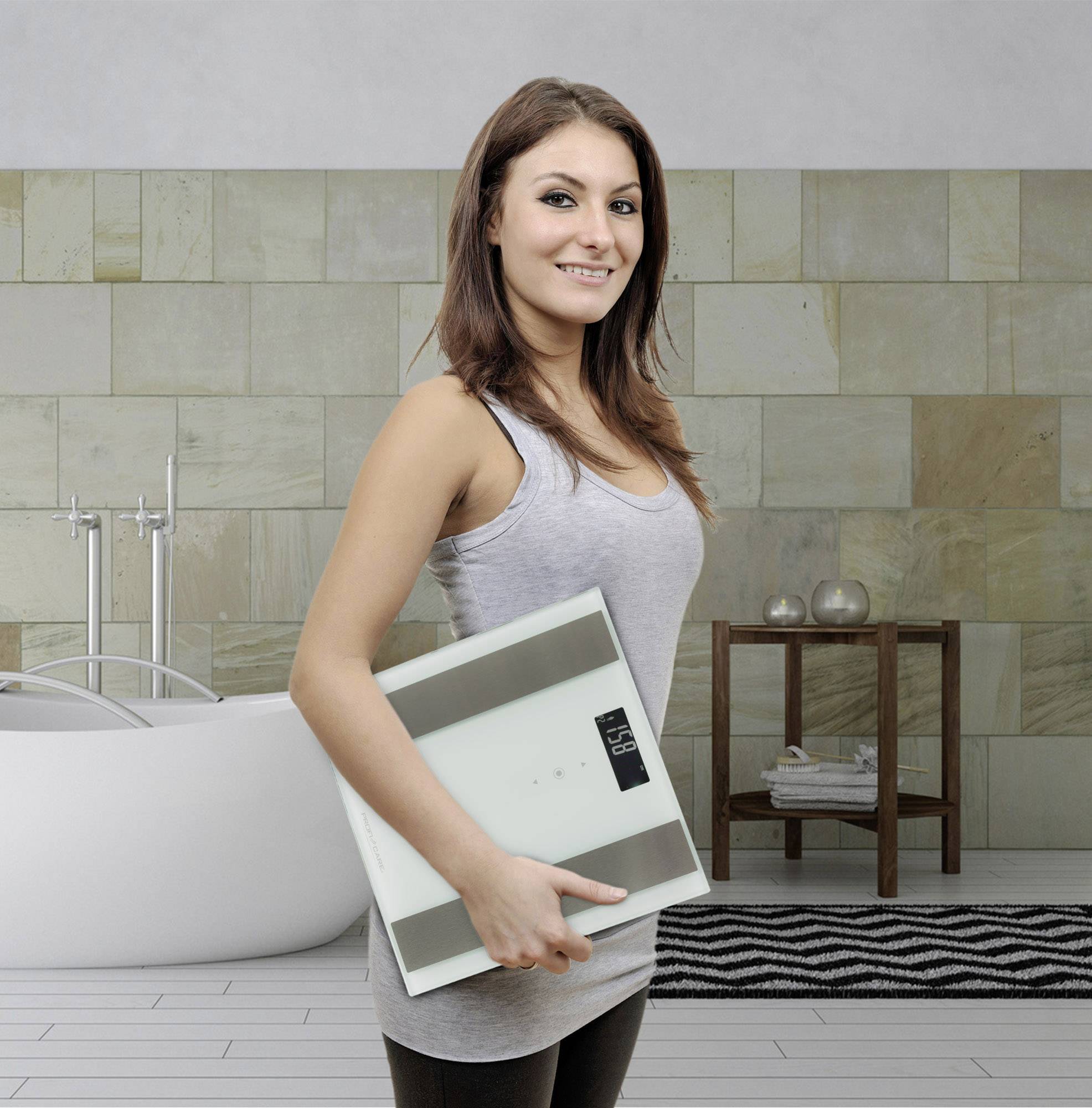 Frau in einem modernen Badezimmer hält eine digitale Personenwaage in den Armen. Hinter ihr ist eine Badewanne und ein Regal zu sehen.