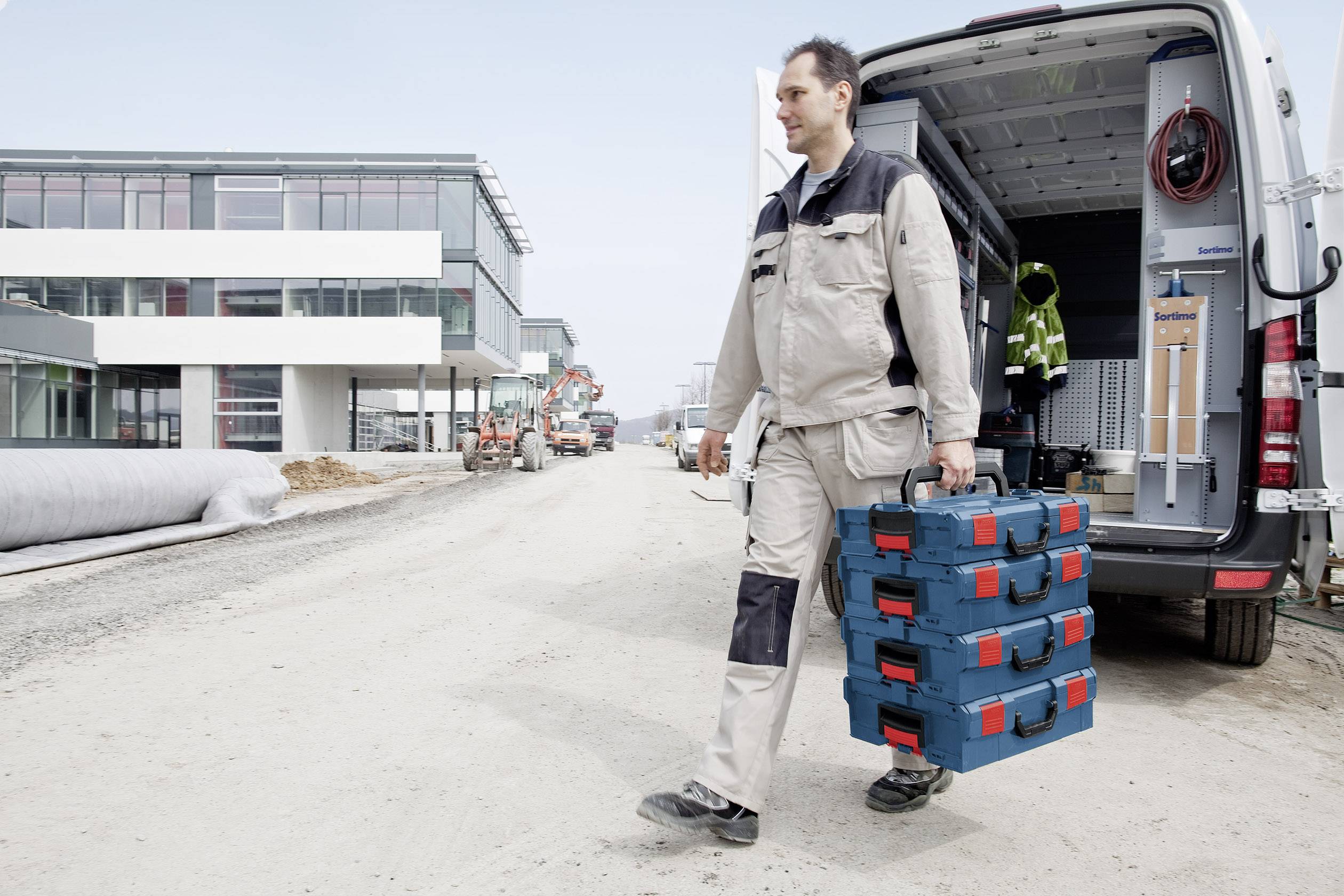 Ein Mann in Arbeitskleidung trägt einen Werkzeugkoffer auf einer Baustelle. Im Hintergrund ist ein Gebäude und ein Lieferwagen zu sehen.