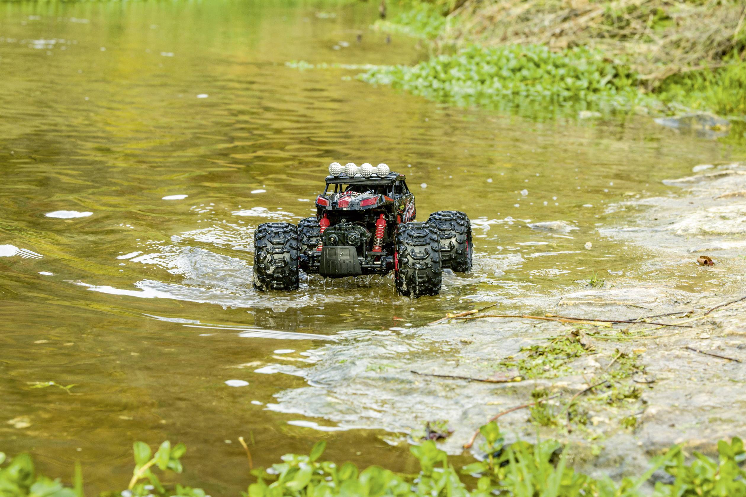 Ein ferngesteuertes Auto mit großen Reifen fährt durch flaches Wasser in einer grünen Umgebung. Es spritzt Wasser auf beiden Seiten.