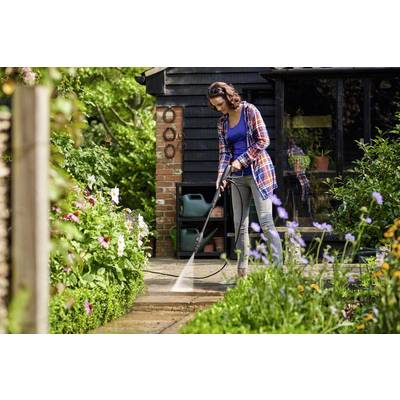 Frau reinigt Terrasse mit Hochdruckreiniger im Garten. Im Hintergrund ein Haus mit Blumen und Pflanzen.