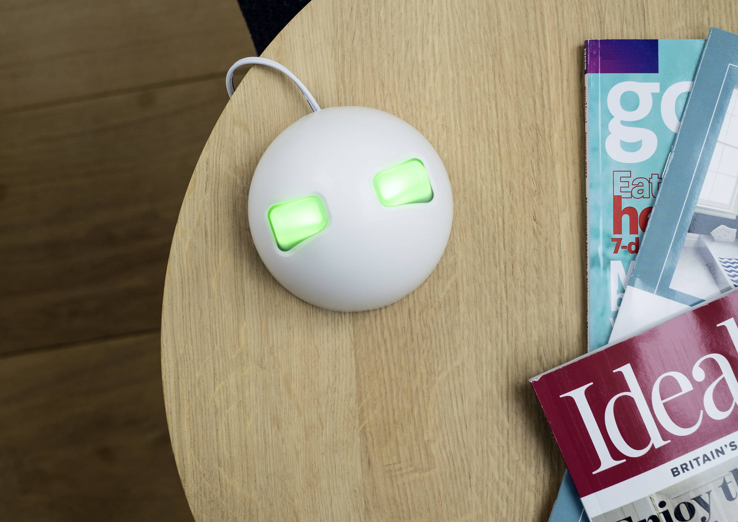 Ein rundes, weißes Gerät mit zwei leuchtend grünen Anzeigen liegt auf einem Tisch neben Zeitschriften. Es ist über ein Kabel verbunden.