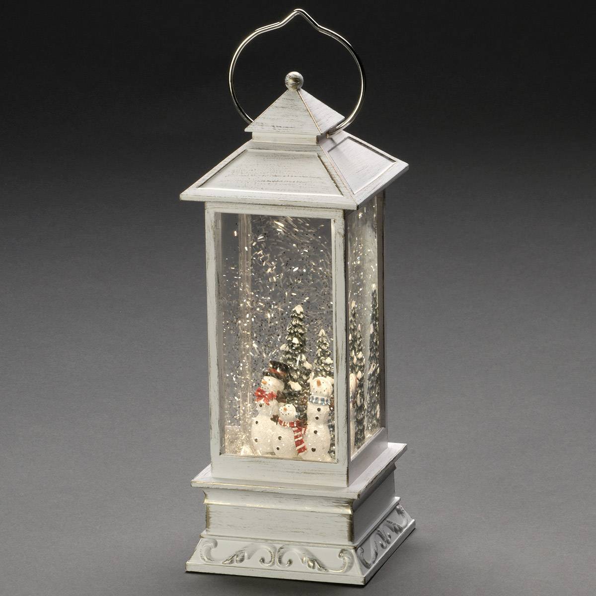 Eine weiße Laterne mit Schneemann- und Baumdekoration. Schneeflocken wirbeln in der Laterne. Sie steht auf einem dunklen Hintergrund.