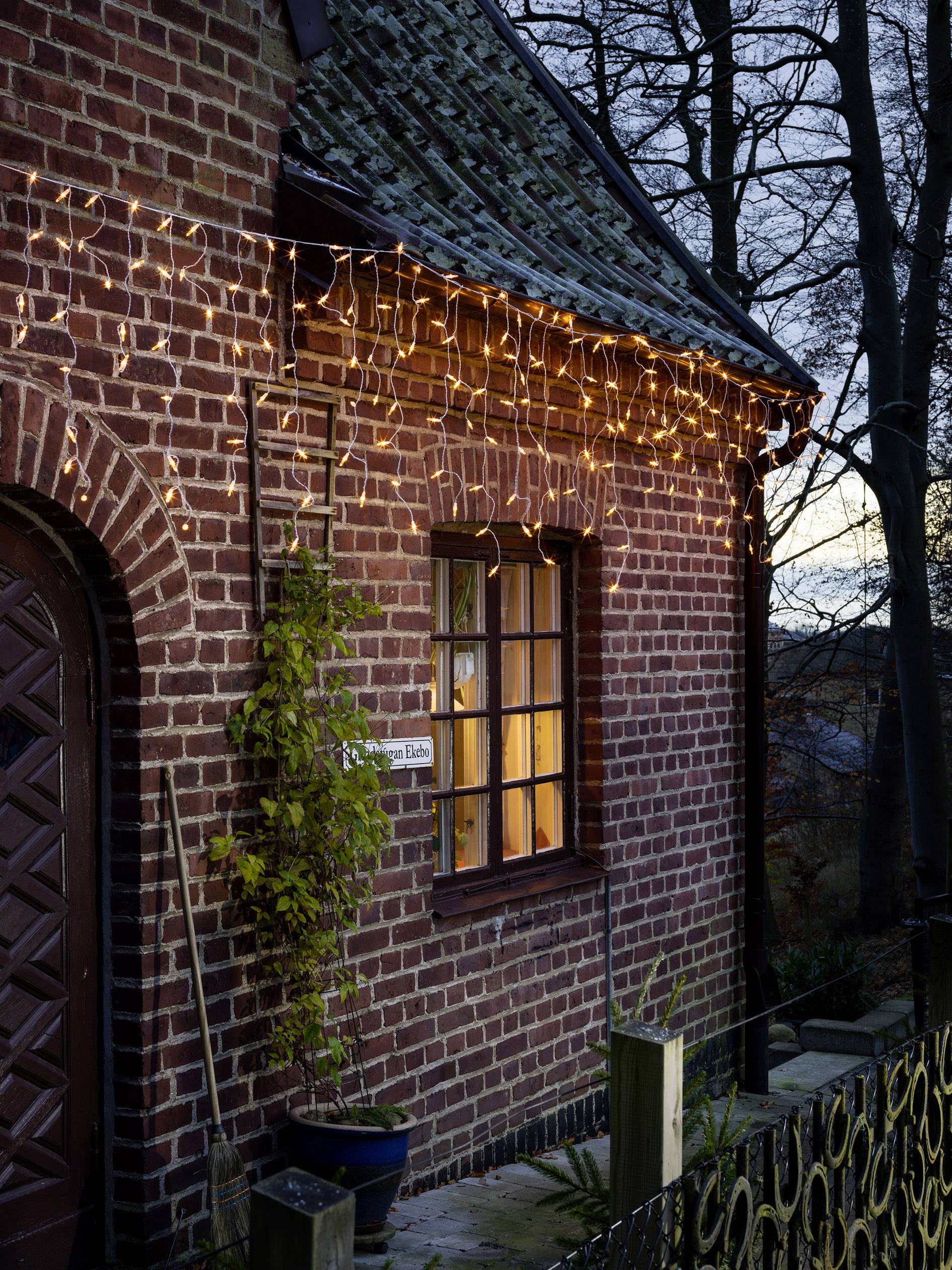 Backstein-Haus mit leuchtender Lichterkette am Abend. Ein warmes Licht scheint aus dem Fenster, Pflanzen daneben an der Fassade.