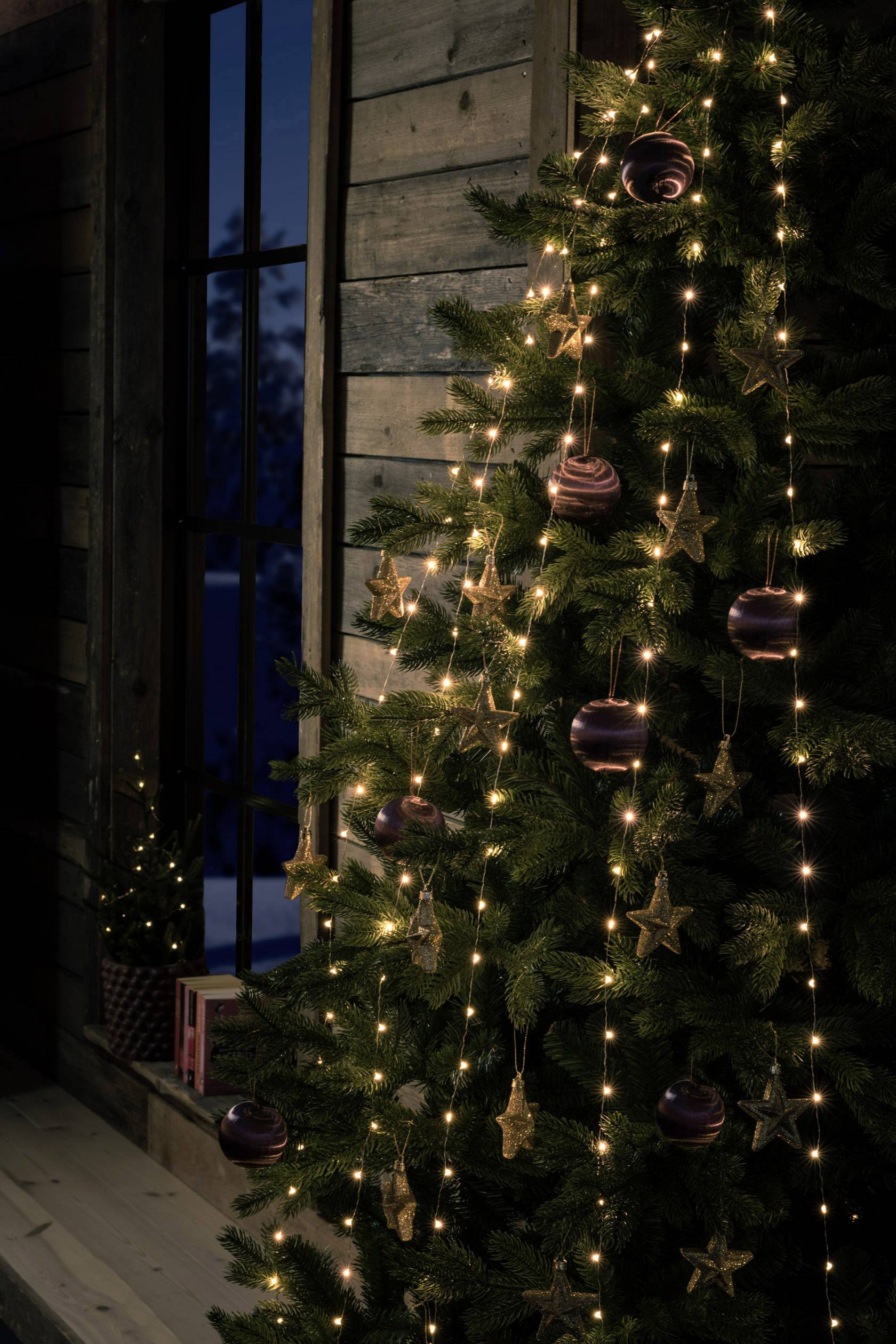 Ein Weihnachtsbaum mit Lichterketten und Schmuckkugeln steht in einem rustikalen Innenraum vor einem Fenster.