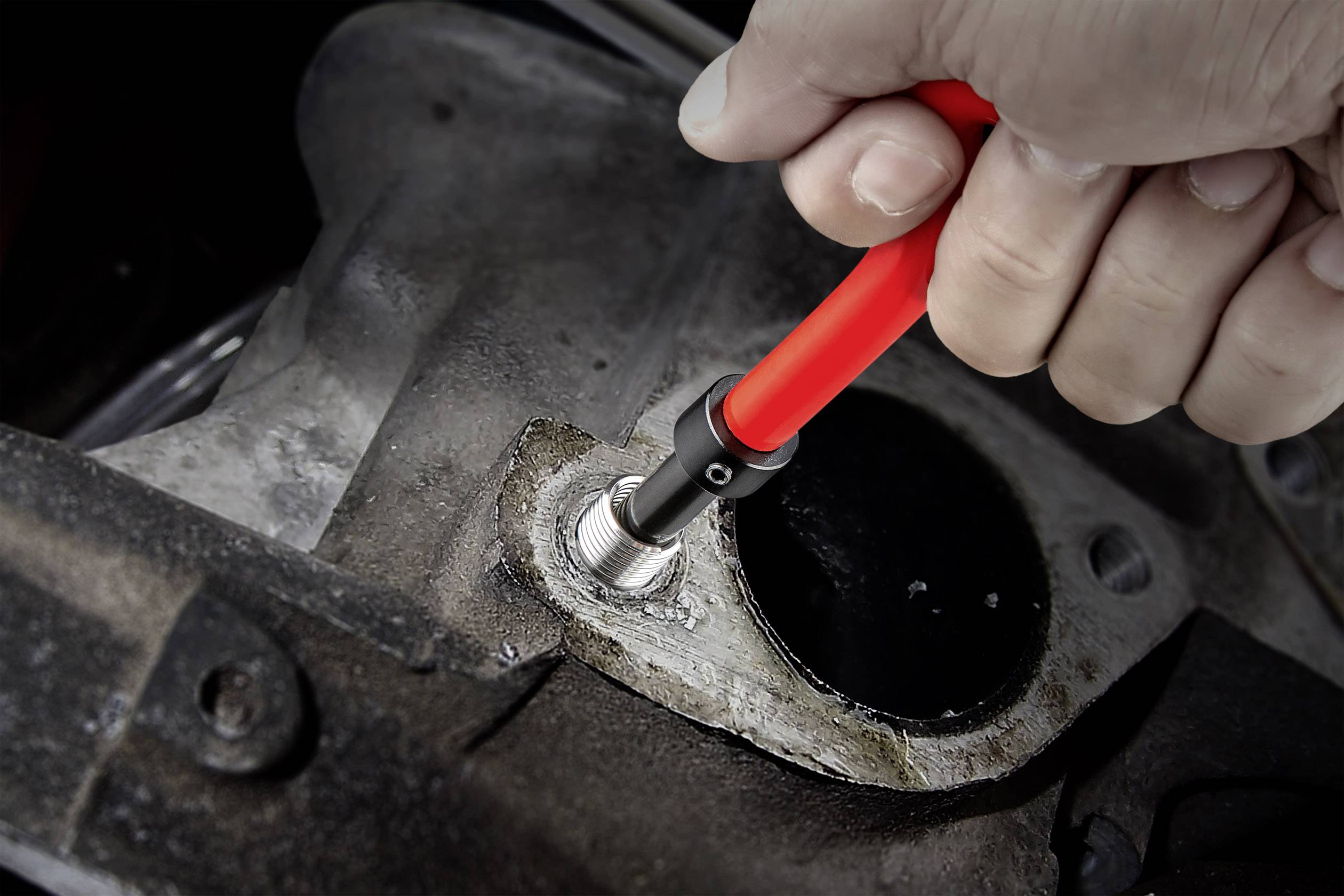 Eine Hand hält ein rotes, flexibles Werkzeug, das eine stark abgenutzte, metallische Schraube an einem grauen, industriellen Gerät festzieht.