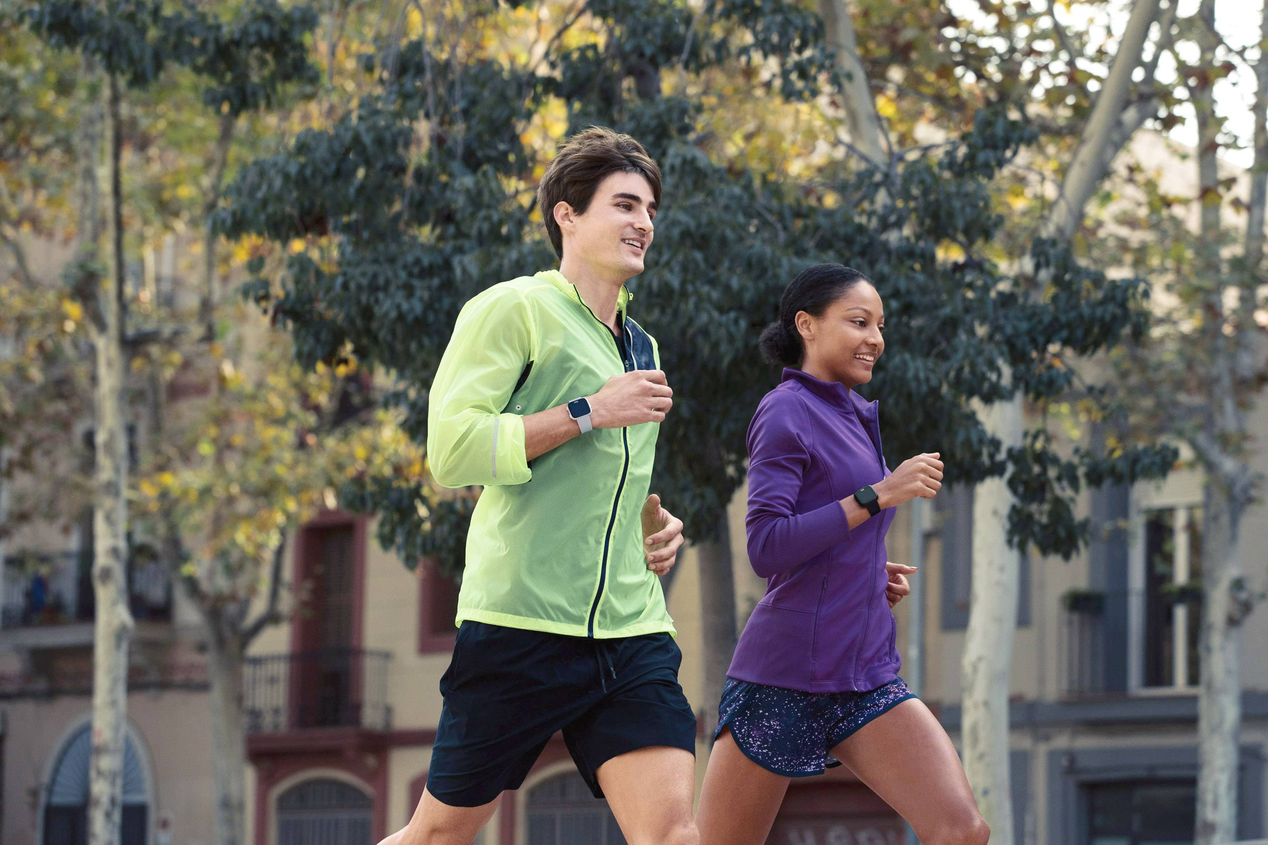 Zwei Personen joggen im Park, mit sportlicher Kleidung und Fitnessuhren. Im Hintergrund sind Bäume und Gebäude zu sehen.