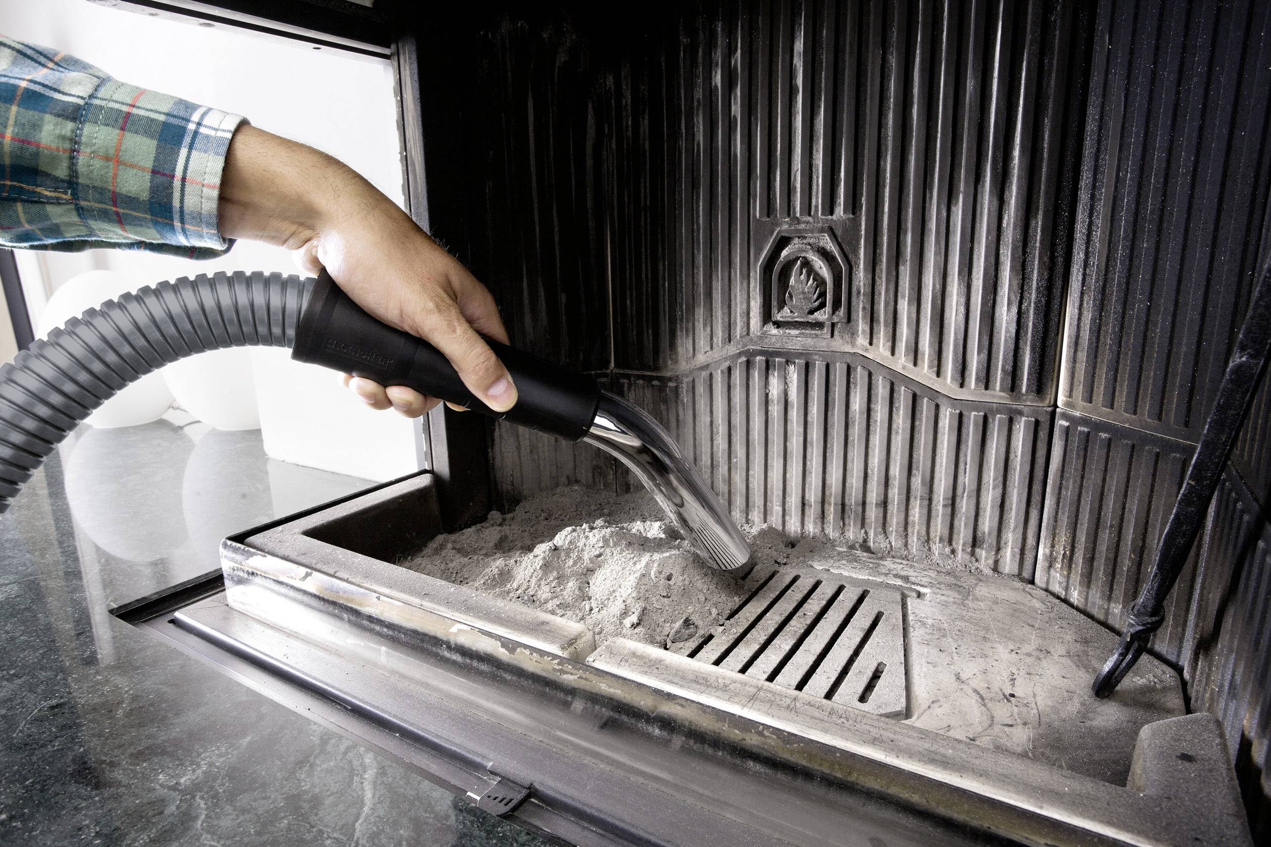Eine Person reinigt einen Kamin mit einem Staubsauger, um Asche zu entfernen. Das Bild zeigt eine Nahaufnahme der Hand und des Staubsaugers.