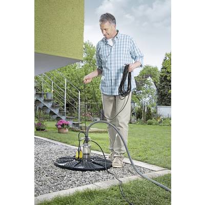 Ein Mann hält ein Kabel neben einer Tauchpumpe im Garten eines Hauses. Im Hintergrund blühen Pflanzen und es führt eine Treppe zum Haus.