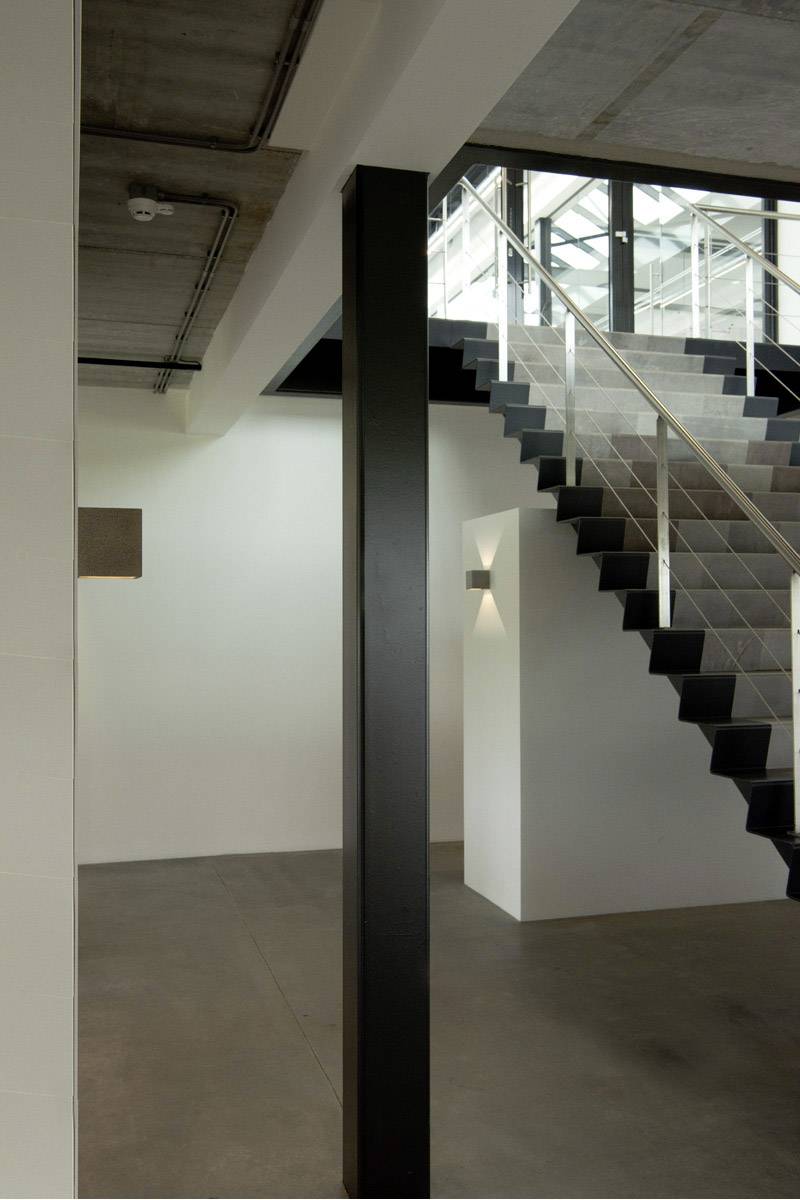 Modernes Treppenhaus mit Stahlgeländer und Betonwänden, industrieller Stil, links ein schwarzer Pfeiler, helle Beleuchtung.