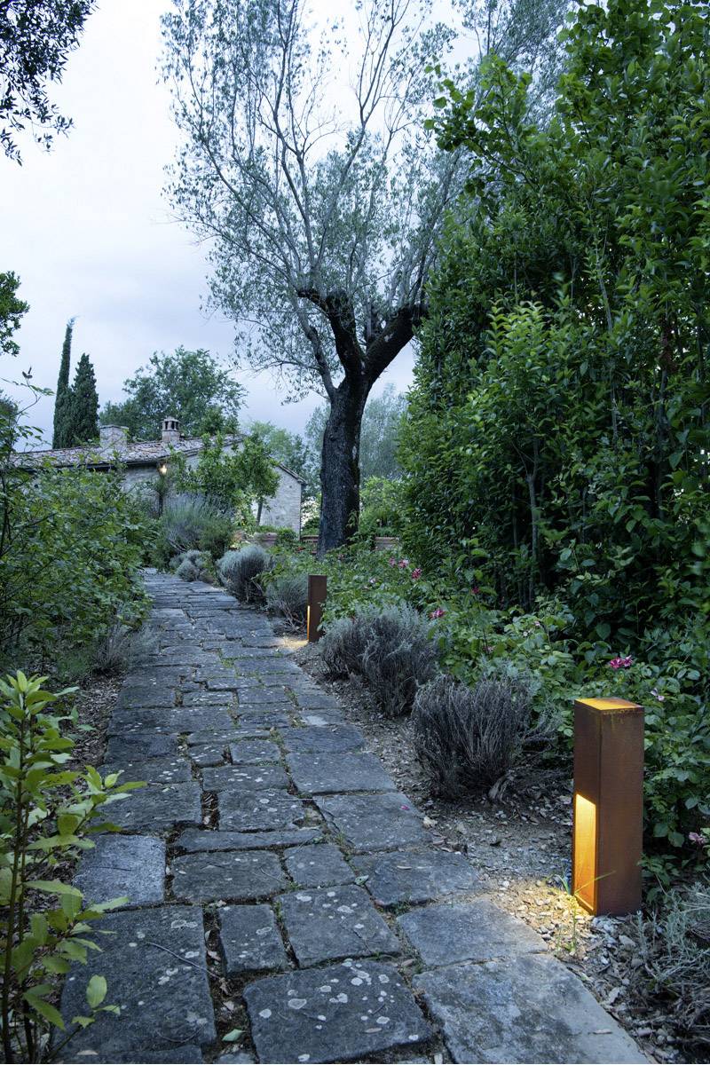 Ein gepflasterter Gartenweg führt durch üppiges grünes Laub zu einem Steinhaus mit Baum im Hintergrund; Beleuchtung entlang des Weges.