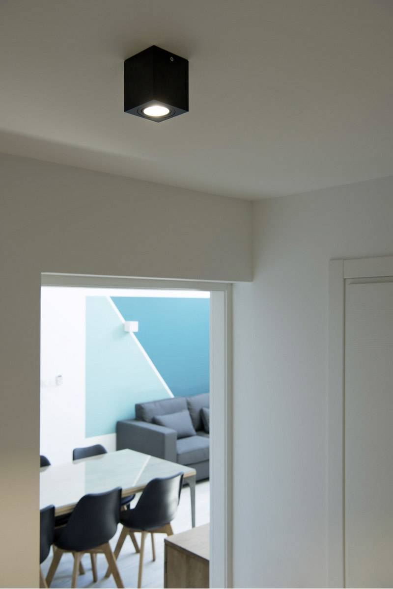 Esszimmer mit modernem Tisch und Stühlen, im Hintergrund ein Sofa und türkisfarbene Wand. Deckenleuchte an der weißen Decke.