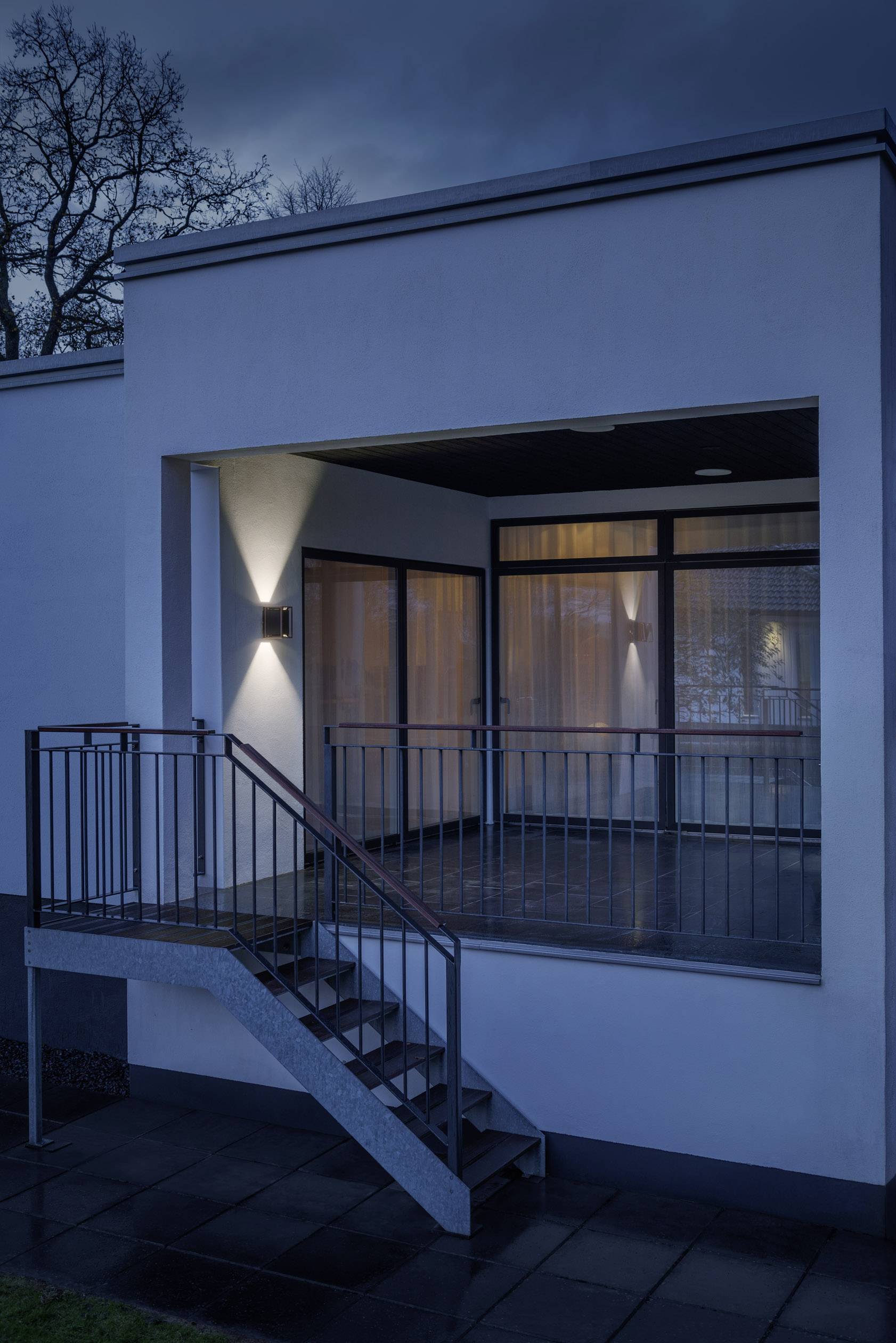 Eine moderne Hausfassade bei Nachtbeleuchtung, zeigt eine überdachte Terrasse mit Metallgeländer und Treppe, umringt von Bäumen.