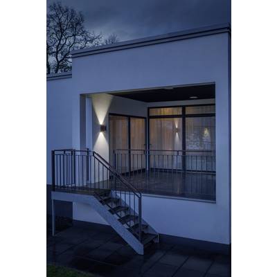 Eine moderne Hausfassade bei Nachtbeleuchtung, zeigt eine überdachte Terrasse mit Metallgeländer und Treppe, umringt von Bäumen.