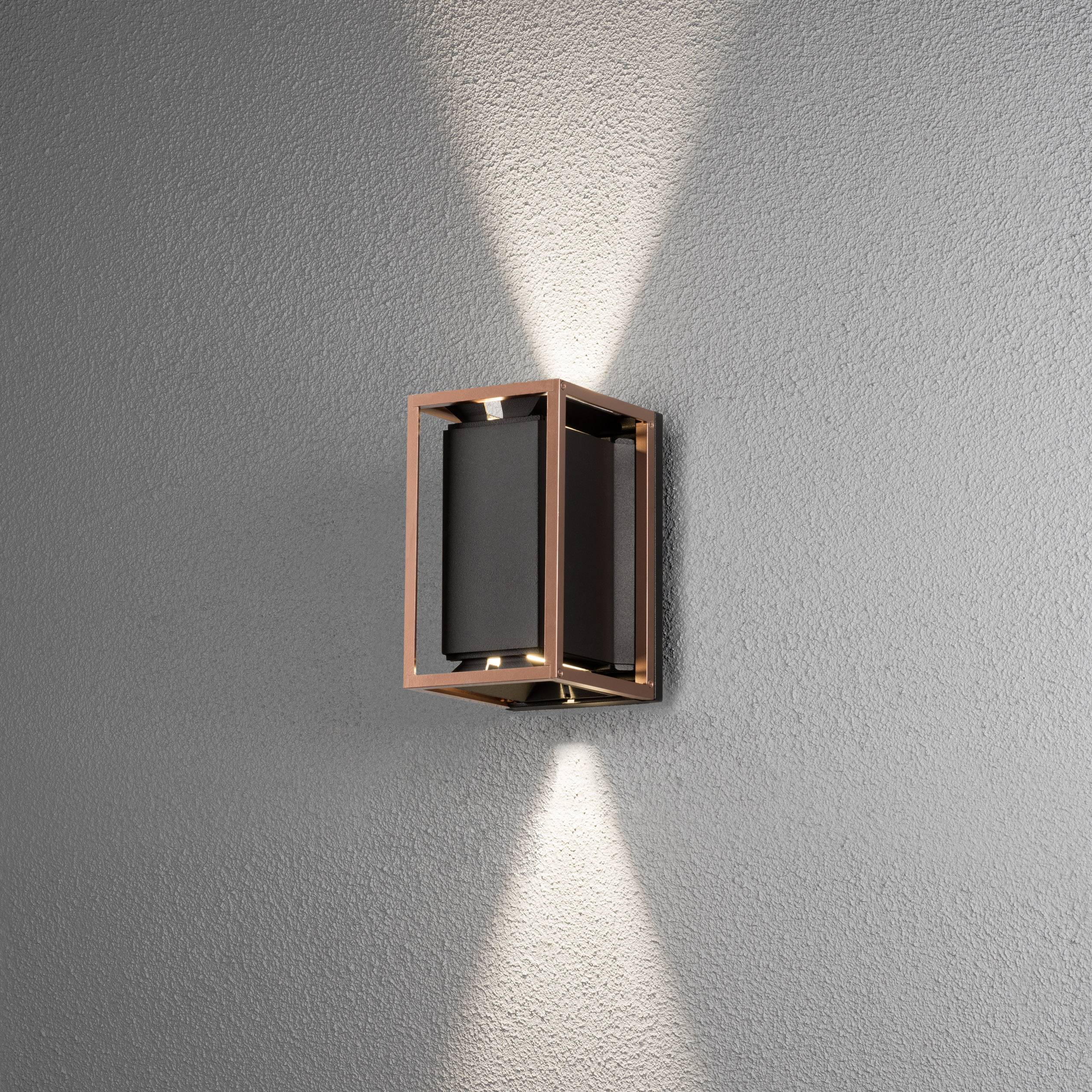 Wandleuchte aus Metall mit quadratischem Rahmen, strahlt Licht nach oben und unten aus, vor grauer Wand montiert.