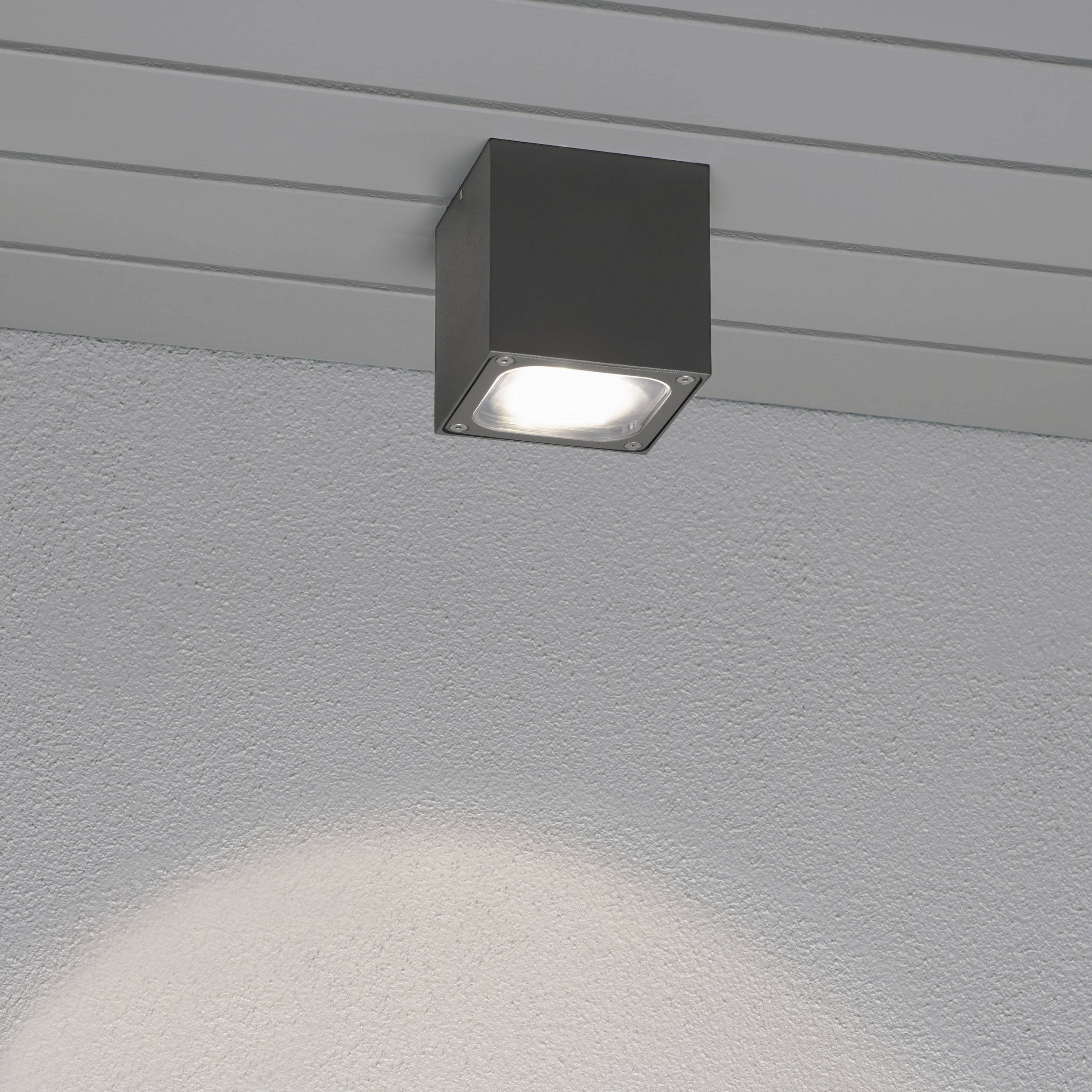 Eine quadratische Deckenlampe leuchtet an einer hellgrauen Decke. Die Lichtquelle erzeugt ein rundes Lichtmuster an der Wand darunter.