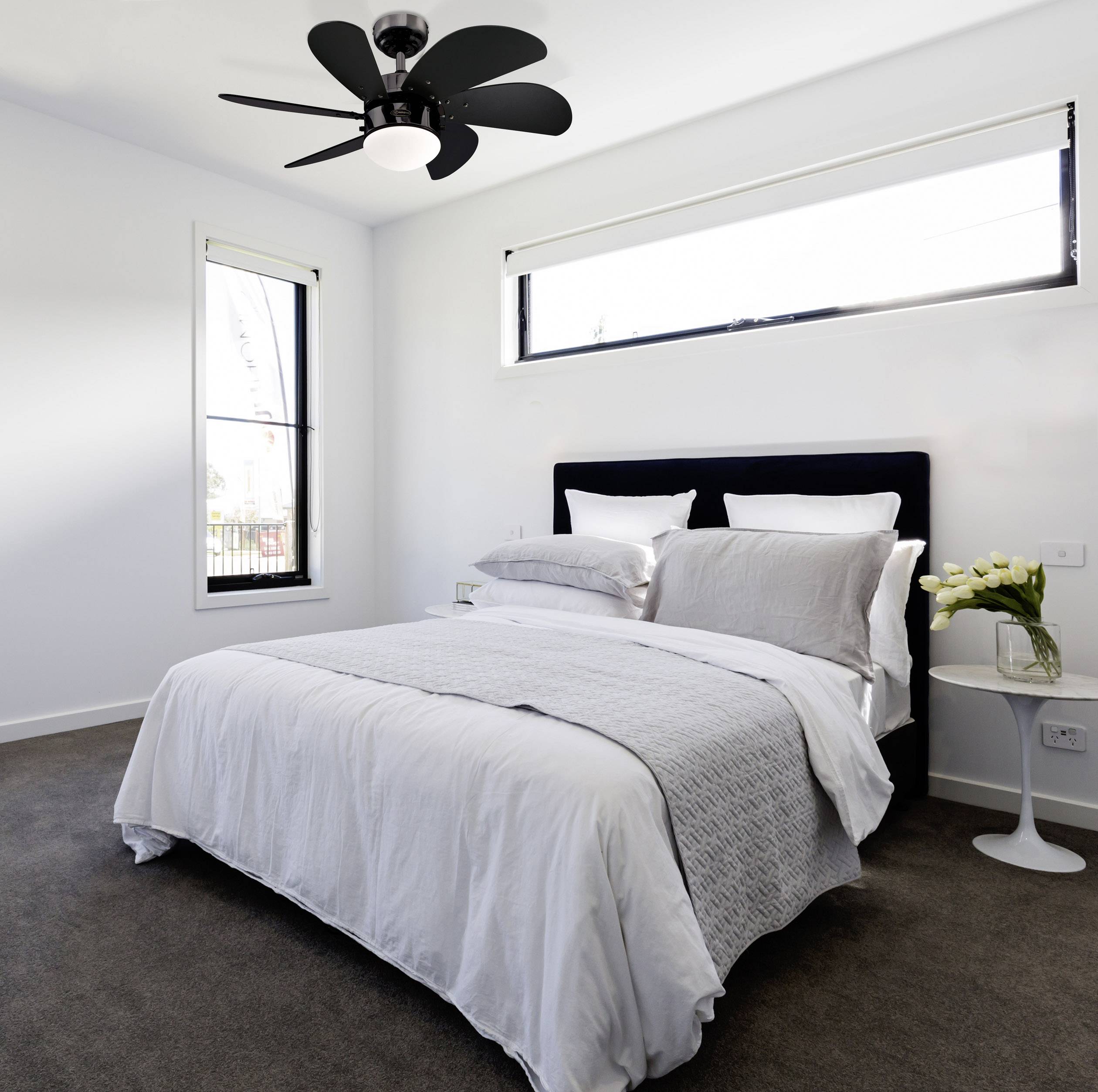 Helles Schlafzimmer mit einem Doppelbett, weißer Bettwäsche, Nachttisch und einem Deckenventilator. Fenster lassen Tageslicht herein.