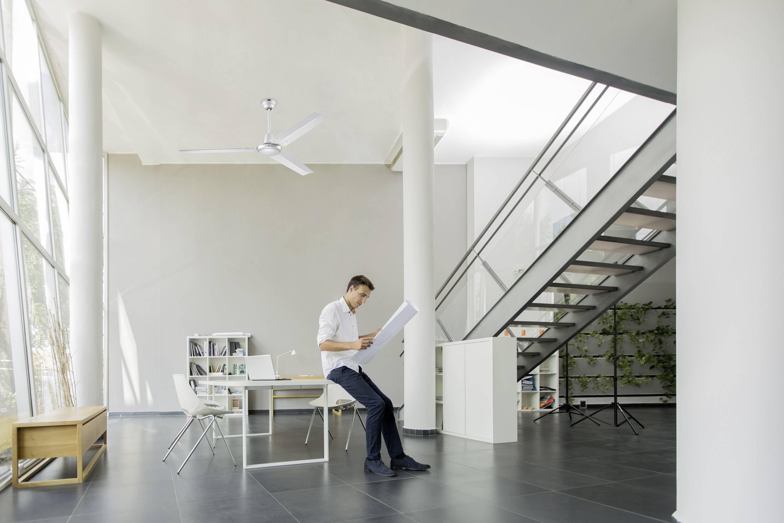 Ein Mann sitzt in einem modernen, hellen Büro und liest ein Dokument. Im Hintergrund sind eine Treppe und große Fenster zu sehen.