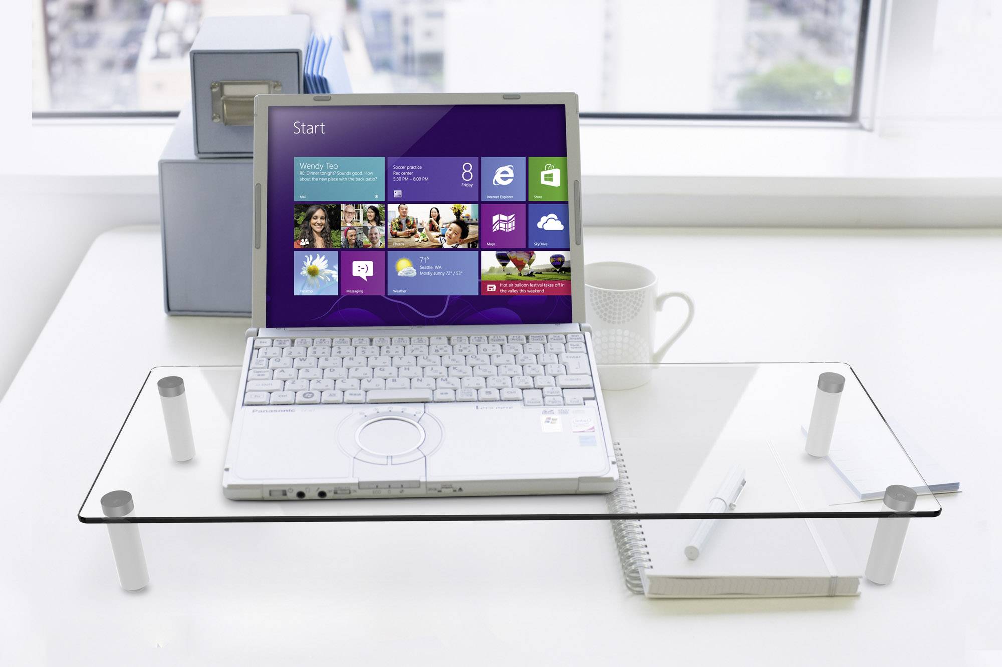 Ein Laptop mit Windows 8 ist auf einem Glastisch platziert. Im Hintergrund ist Fensterblick auf eine Stadt. Daneben stehen eine Tasse und ein Notizbuch.