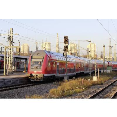 Ein roter Doppelstockzug fährt bei sonnigem Wetter durch einen Bahnhof. Im Hintergrund sind Hochhäuser und Oberleitungen zu sehen.