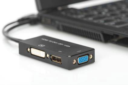 Ein Laptop ist mit einem Adapter verbunden, der VGA-, DVI- und HDMI-Anschlüsse bietet. Der Adapter liegt auf einer weißen Oberfläche.