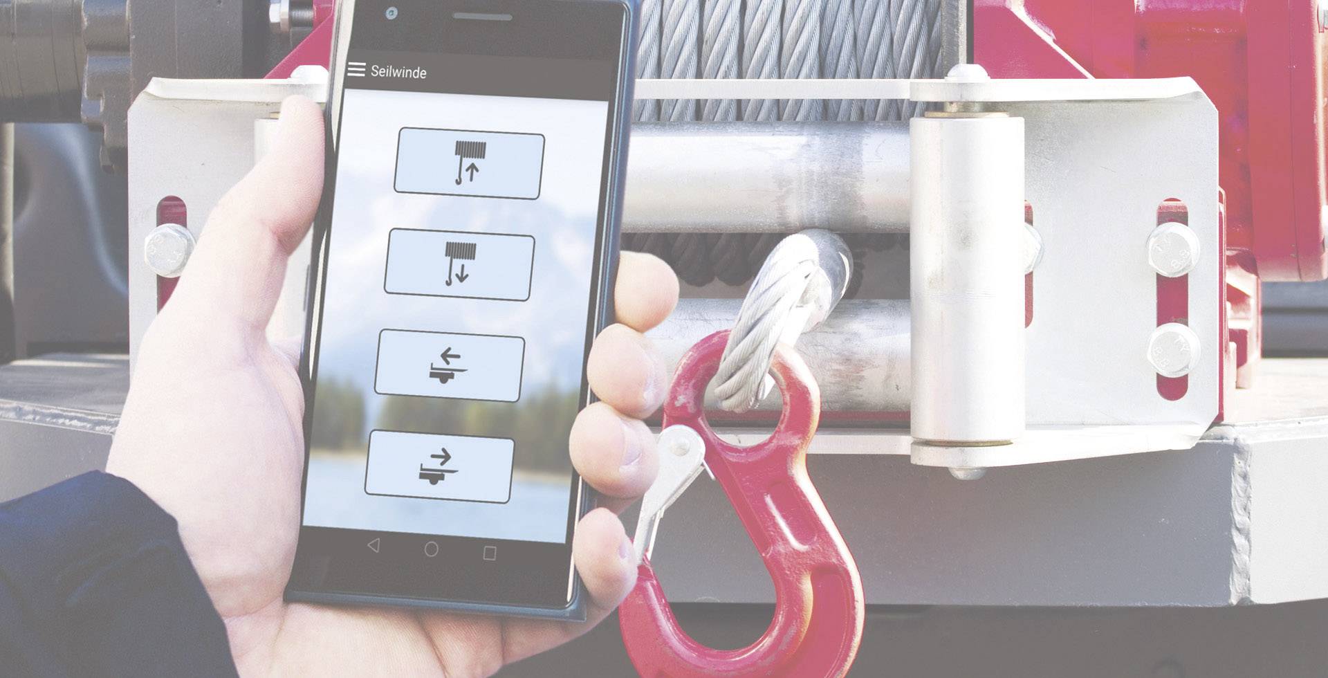 'Eine Hand hält ein Smartphone mit einer App zur Steuerung einer Seilwinde. Im Hintergrund ist ein Teil der Winde sichtbar.'