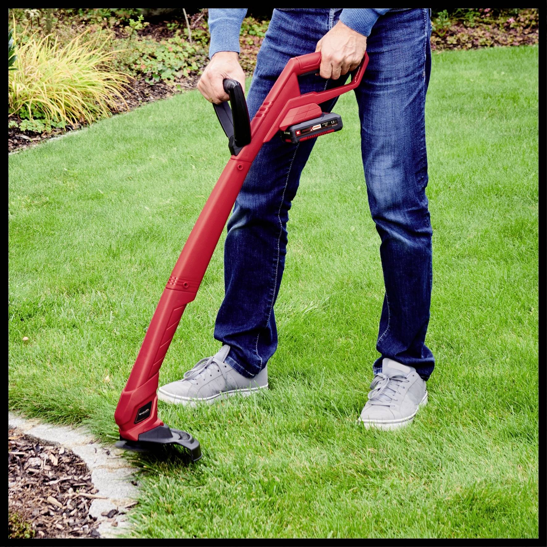 Eine Person verwendet einen roten elektrischen Rasentrimmer, um den Rand eines grünen Rasens zu schneiden. Im Hintergrund sind Pflanzen zu sehen.