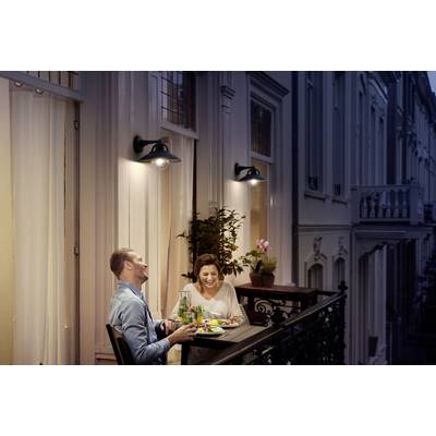 Ein Paar sitzt auf einem Balkon, isst zu Abend und lacht. Die Atmosphäre ist gemütlich, mit warmem Licht und einer Pflanze im Hintergrund.