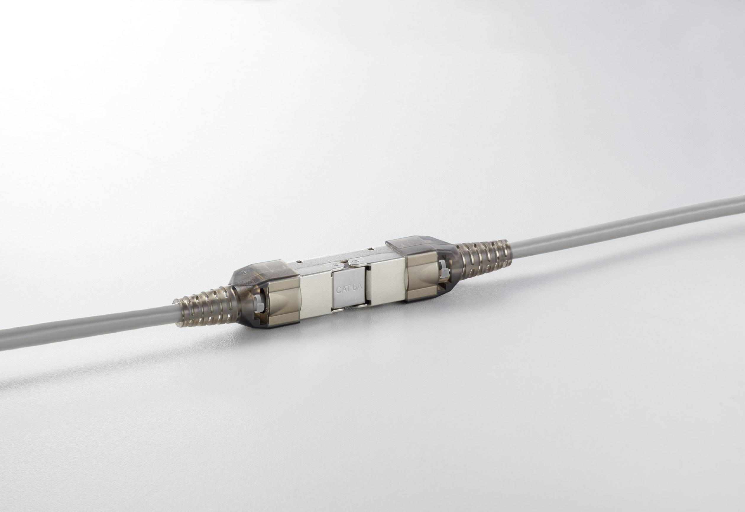 Ein graues Kabel mit einem metallischen Verbinder in der Mitte liegt auf einer weißen Oberfläche. Der Verbinder hat eine rechteckige Form.