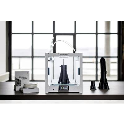 Ein 3D-Drucker druckt ein schwarzes Objekt. Links ist eine Box mit Filament, rechts fertige Objekte. Fenster im Hintergrund.
