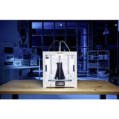 Ein moderner 3D-Drucker auf einem Tisch druckt ein schwarzes Objekt. Im Hintergrund sind Regale mit Modellprototypen sichtbar.