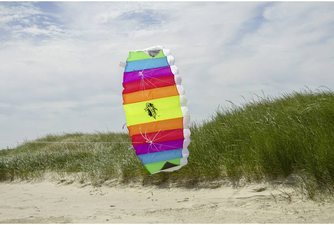Ein bunter Drachen fliegt knapp über den Sanddünen eines Strandes unter einem bewölkten Himmel.